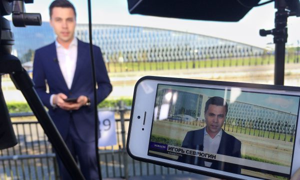 A mobile phone shows a reporter outdoors, the broadcast setup is out of focus in the background