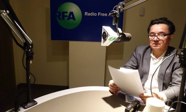 A man reads into a microphone, RFA logo in the background