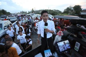 Reporter with RFA mic speaks to a camera. He is on the back of a flat bed truck, in a procession. Most people are dressed in white.