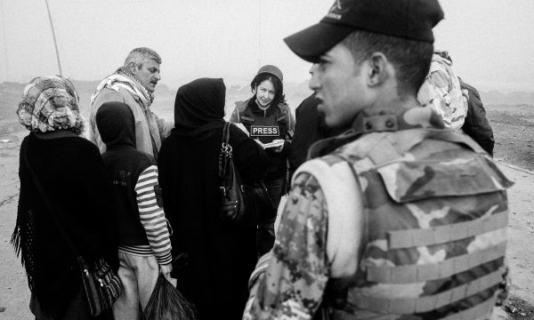 a woman wearing a press flak jacket listens to people who have surrounded her