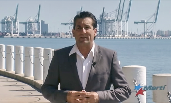 a middle-aged man in a gray suit jacket and no tie stands near a bay and large cranes in the distance