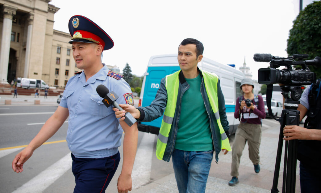 People walking down the street: an officer, followed by a journalist with a microphone, camera being held by someone out of frame and a woman with a photo camera further back.